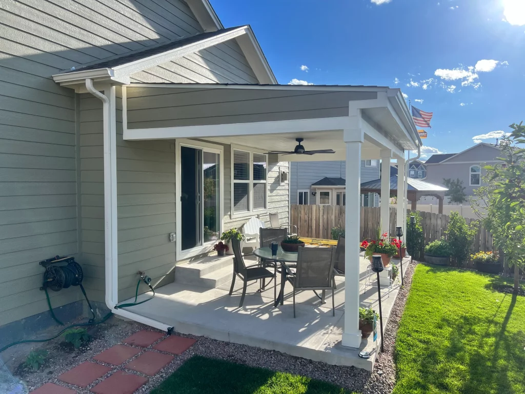 New covered patio with white posts and ceiling fan attached to modern house with green lawn and potted flowers.